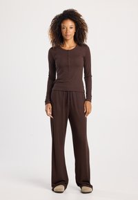 Ruskea pitkähihaisten paita, jossa nappikoristeita, yhdistettyinä löysiin ruskeisiin housuihin. Molemmat ovat valmistettu pehmeästä ja sileästä kankaasta.