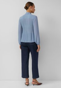 Hellblaue Bluse mit plissiertem Rücken, langen Ärmeln und Kragen, kombiniert mit dunkelblauen, weit geschnittenen Jeanshosen und braunen Sandalen mit Absatz.