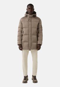 Puffer jacket in beige, featuring a high collar, hood with drawstrings, zippered pockets, and a quilted design. Paired with cream pants and brown sneakers.