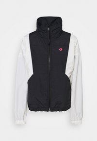 Veste légère avec manches longues blanches, devant et dos matelassés noirs, fermeture éclair à l'avant, et petit logo rose sur la poitrine.