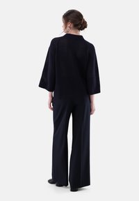 Frau mit einem Dutt, die eine marineblaue, weite Hose und einen locker geschnittenen marineblauen Pullover trägt, steht mit dem Rücken vor einem weißen Hintergrund.