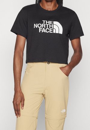 Osoba nosząca czarny krótki t-shirt z logo "THE NORTH FACE" i beżowe spodnie z przednimi kieszeniami, jedną ręką w kieszeni.