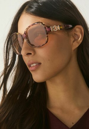 Kobieta z długimi ciemnymi włosami nosząca duże okulary przeciwsłoneczne w oprawkach typu żółw z złotymi dekoracyjnymi akcentami na zausznikach, patrząca lekko w prawo.