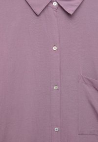 Chemise à boutonnières de couleur violet clair en tissu texturé, avec un col, six boutons et une poche poitrine sur le côté gauche.