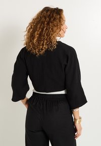 Top corto negro con mangas anchas, hecho de una tela texturizada. Se lleva con pantalones negros de cintura alta. Diseño minimalista, acentos de estilo sutiles.