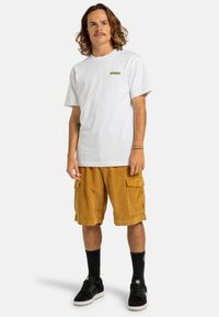 Hvid bomulds t-shirt med et lille grønt logo, parret med brune kordfløjls cargo shorts med to sidelommer, sorte sneakers og sorte sokker.