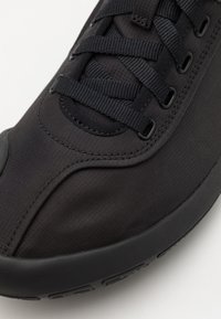 Black athletic shoe with a smooth synthetic upper, textured fabric, and flat black laces. Round toe and cushioned rubber sole.