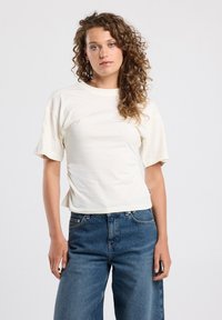 Cream-colored, short-sleeve t-shirt with a relaxed fit, featuring side ruching and a round neckline, paired with high-waisted denim jeans.