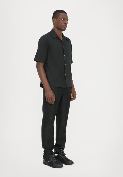 Chemise noire avec un motif texturé, à manches courtes et boutons dorés, assortie à un pantalon noir texturé et des chaussures de sport noires.
