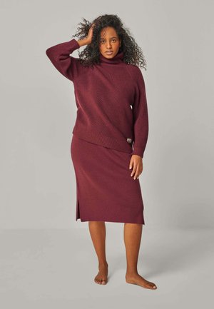 Frau mit lockigem Haar, die einen burgunderroten Strickrollkragenpullover und einen passenden Midirock trägt, steht barfuß vor einem schlichten Hintergrund.