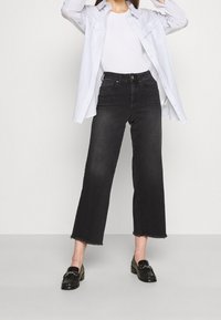 Černé kalhoty s širokými nohavicemi a roztrhaným lemem, doplněné bílým tričkem a světlomodrou pruhovanou košilí. Celkový vzhled doplňují černé mokasíny.