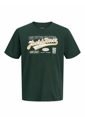 Teenage dreng står iført en mørkegrøn grafisk t-shirt, løse brune bukser og beige sneakers på en ensfarvet hvid baggrund.