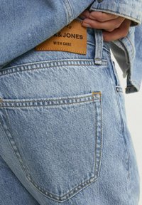 Jeans din denim albastru deschis cu un finisaj texturat, având un eticheta din piele cu litere embossate și cusături portocalii contrastante.