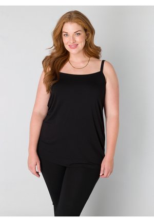 Lächelnde Frau mit langen roten Haaren, die ein schwarzes ärmelloses Oberteil und schwarze Hosen vor einem schlichten hellen Hintergrund trägt.