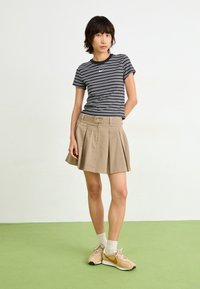 Ung kvinna iklädd en svart- och vitrandig kortärmad topp, beige plisserad kjol, vita strumpor och bruna Nike-sneakers står på en grön golv.