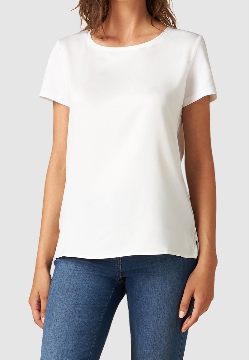 Femme portant un chemisier blanc uni à manches courtes et un jean bleu, debout devant un fond gris clair.