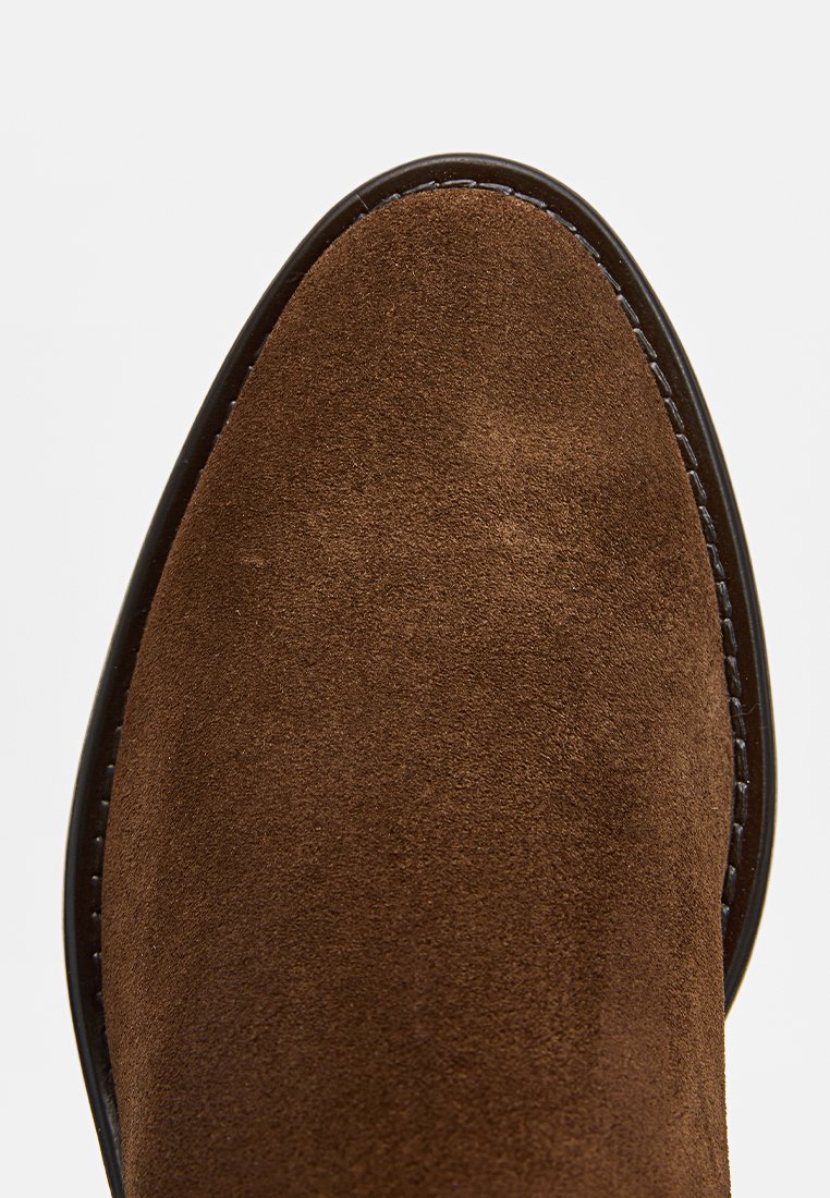 Botín de ante marrón hasta el tobillo con textura suave, punta redondeada y costuras contrastantes a lo largo del lateral. Diseño clásico con una elegancia sutil.