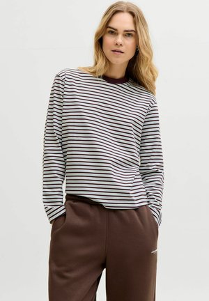 Frau mit blondem Haar, die ein hellblau-braun gestreiftes Langarmshirt und braune Hosen trägt, mit den Händen in den Taschen, nach vorn blickend.