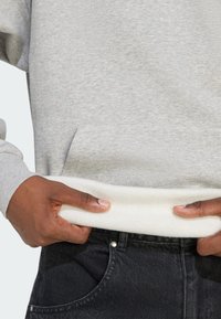 Sivá mikina s bielym lemom obloženým flísom, vybavená predným vreckom. Ruky spočívajúce na vrecku, zobrazujúce kontrast textúry látky.