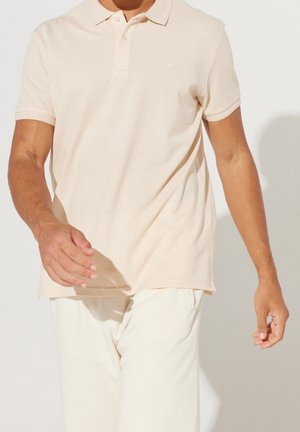 Mann in einem hellbeigen Polohemd mit kurzen Ärmeln und cremefarbenen Hosen, der mit entspannten Armen neben seinem Körper geht.