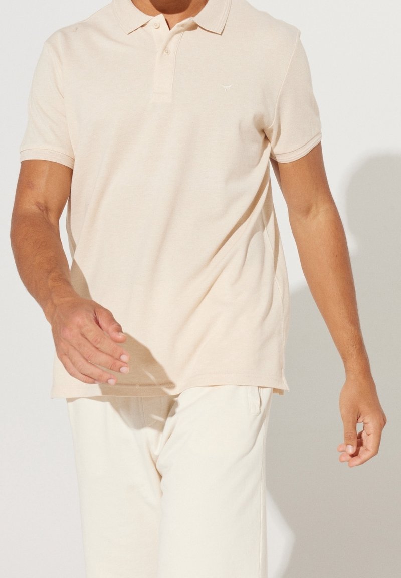 Hombre vistiendo una camiseta polo de manga corta beige claro y pantalones color blanco roto, caminando con los brazos relajados a los lados.