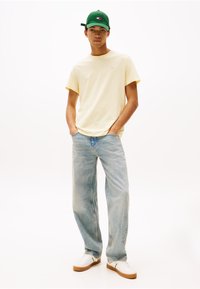 T-shirt en coton jaune clair, coupe décontractée ; jean en denim bleu clair, coupe large ; casquette verte avec logo ; baskets blanches avec semelle en caoutchouc.