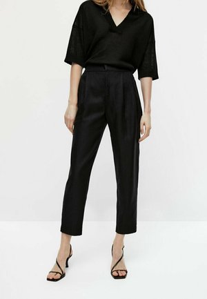 Kvinde stående iført sort V-hals kortærmet top, sorte skræddersyede bukser med høj talje og sorte sandaler med hæl og stropper mod hvid baggrund.