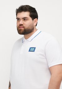 camiseta polo blanca con ribete azul en el cuello y un logo cuadrado azul en el lado izquierdo del pecho, con mangas cortas y un corte clásico.
