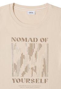 T-shirt beige avec texte métallique "NOMAD OF YOURSELF" et graphique rectangulaire rayé en tons or et argent.