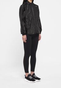 Kobieta w czarnej lekkiej kurtce z kapturem, czarnych legginsach z siateczkowymi panelami i czarno-białych sneakersach, stojąca na białym tle.