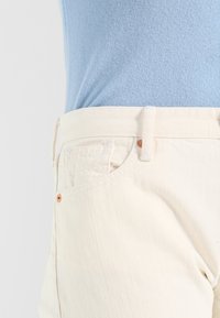 Los pantalones color crema presentan una textura tejida, un diseño estándar de cinco bolsillos y un sutil botón de acento naranja en el frente.