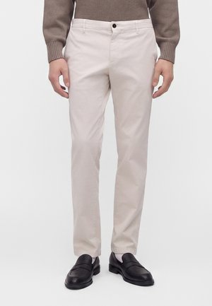 Mann trägt beige Hose, braunen Pullover, weiße Socken und schwarze Leder-Slipper, steht vor einem einfarbig weißen Hintergrund.