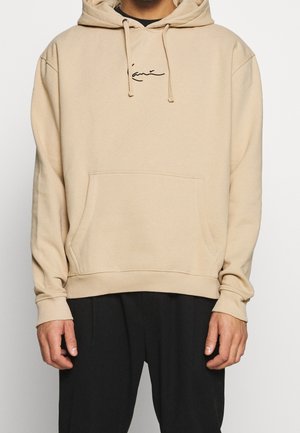 Sudadera beige fabricada con un tejido suave, que presenta un bolsillo delantero tipo canguro, capucha con cordón y un sutil logo negro bordado en el pecho.