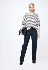 Sweat-shirt blanc et noir à rayures, surdimensionné, jean large bleu foncé, sac à main noir et chaussures noires épaisses. Le mannequin se tient devant un fond clair.