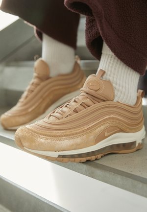 Beige Nike Air Max 97 sneakers gedragen met witte sokken en bruine broek, rustend op een betonnen trapstap.