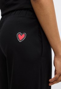 Pantaloni negri dintr-un material moale, cu talie elastică și un desen în formă de inimă, roșu și alb, pe zona buzunarului din spate.