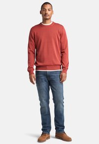 Rustkleurige gebreide trui met een ronde hals, gedragen over een witte blouse, gecombineerd met blauwe jeans en tan schoenen. Regular fit en lichte ribbels bij de manchetten.