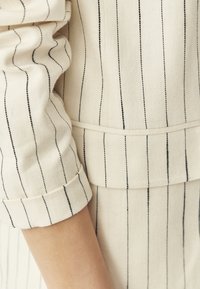Béžová látka s černými vertikálními pruhy, s ozdobným manžetou a detailem švu. Textura vypadá tkaná a mírně hrubá.