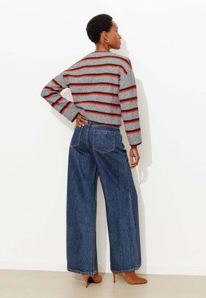 Pull rayé gris avec des lignes rouges et noires, associé à un jean denim bleu à jambes larges. La tenue est complétée par des chaussures à talons marron.