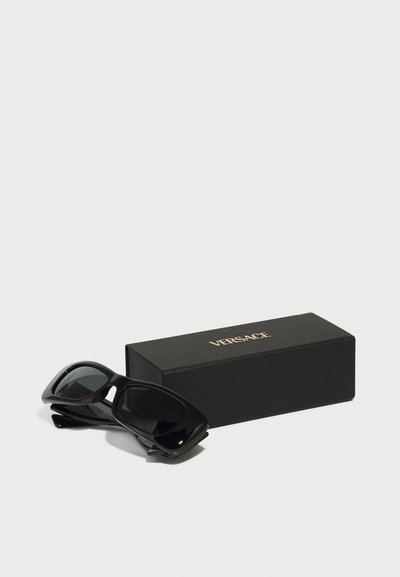 Melni saulesbrilles ar spīdīgu plastmasas rāmi un tumšiem stikliem, novietotas blakus melnai kastītei ar zeltītu "VERSACE" zīmolu.