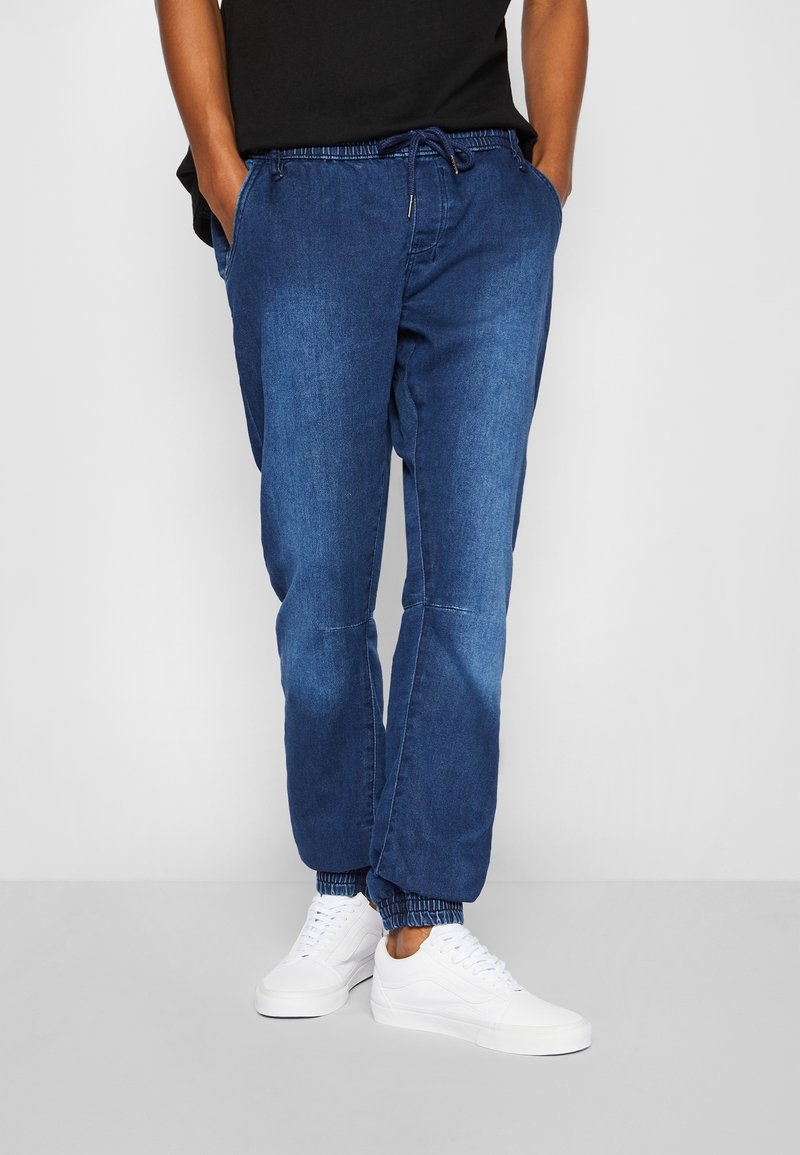 Personne portant un pantalon jogger en denim bleu avec des poignets élastiques et des baskets blanches, mains dans les poches, sur un fond clair uni.