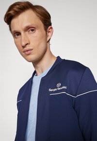 Tumši zila sporta jaka ar baltām akcentu svītrām, priekšējais zips un logo uz kreisā krūšu rajona, uzvilkta virs gaiši zila T-kreklu.