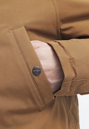 Hand gedeeltelijk in de voorkantzak van een bruin Carhartt-jack, waarbij de textuur van de stof en het metalen drukknoopdetail zichtbaar zijn.