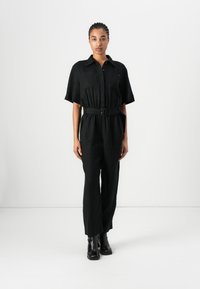 Mono de lino negro con mangas cortas, cintura con cinturón y dos bolsillos en el pecho; combinado con botas negras hasta el tobillo.