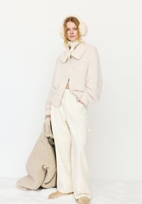 Vrouw in een crèmekleurige, pluizige jas, wijde broek, oorwarmers en sjaal, die een bijpassende oversized jas vasthoudt tegen een witte achtergrond.