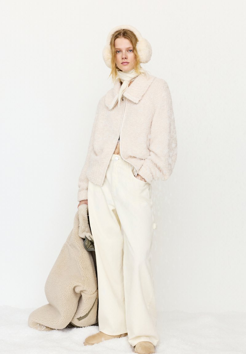 Vrouw in een crèmekleurige, pluizige jas, wijde broek, oorwarmers en sjaal, die een bijpassende oversized jas vasthoudt tegen een witte achtergrond.