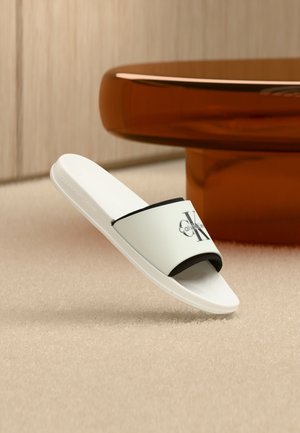 Weiße Pantoletten mit schwarzem Calvin Klein Logo auf dem Riemen, auf beigem Teppich neben einem runden braunen Tisch platziert.