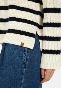 Sweter wykonany na drutach, z kremowymi i granatowymi poziomymi paskami, prążkowanymi mankietami, bocznymi rozcięciami oraz małą tanowymi naszywką z logo blisko hemu.