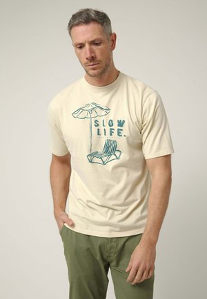Hombre con camiseta beige con texto azul "SLOW LIFE" y gráfico de silla de playa y sombrilla, combinado con pantalones verde oliva.