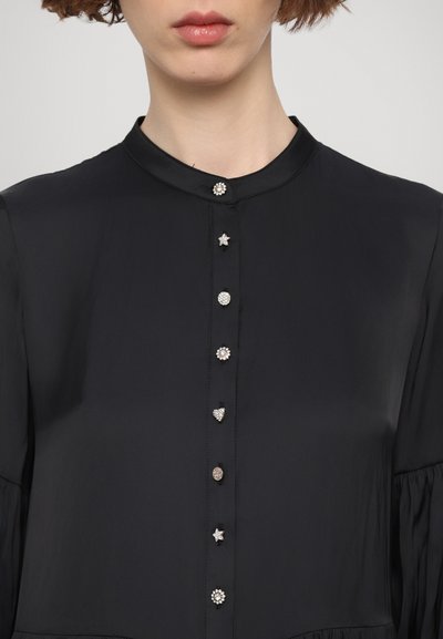 Zwarte blouse met een hoge kraag, voorzien van een rij decoratieve knopen in de vorm van sterren, harten en edelstenen. Glad textuur van de stof.